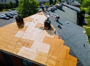 A roofing crew installing asphalt shingles on a two-story home with trees and ladders nearby.